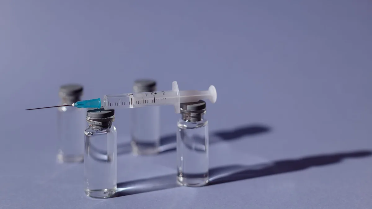 A syringe balanced on vaccine vials with a purple backdrop.